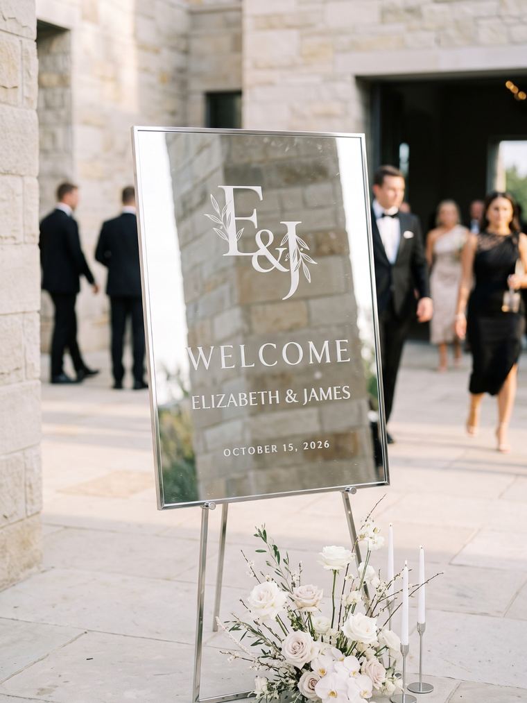 Silver-framed engraved mirror welcome sign with a monogram, greeting guests at a stone venue entrance
